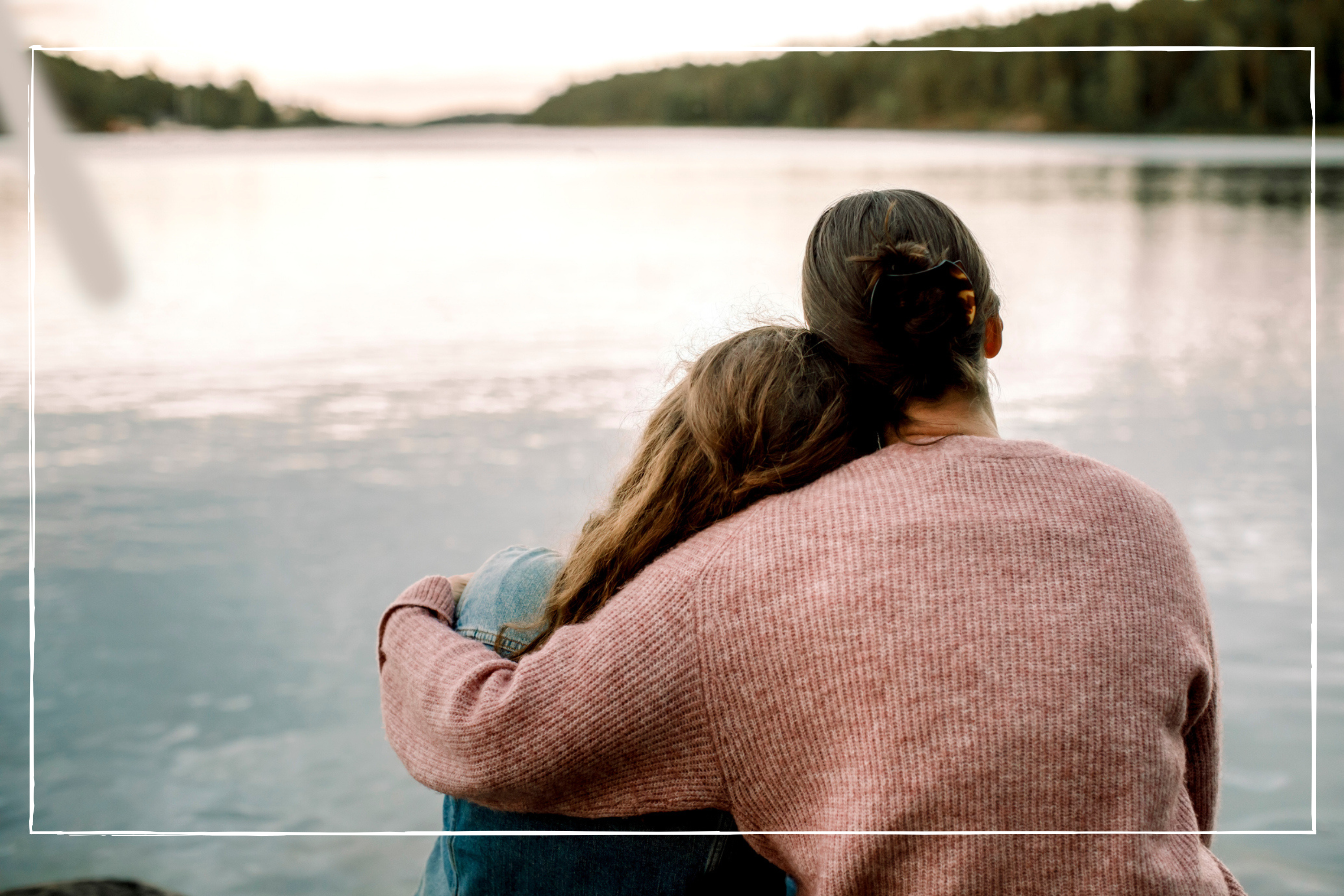 A mother with her arm around her daughter, who is leaning on her shoulder - they're looking out onto a pond 